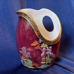 May include: A maroon ceramic pitcher with a unique handle design. The rim and handle are white with gold trim and black detailing. The pitcher features a colorful Asian-inspired landscape design with trees, pagodas, and bridges.