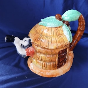 May include: A ceramic teapot shaped like a thatched-roof hut with a palm tree handle and spout. The teapot features a brown, tan, and white color scheme with a red accent. The lid is adorned with green leaves and brown coconuts.