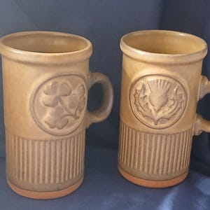 May include: Two tan ceramic mugs with handles. One mug features a raised shamrock design, the other a thistle. Both mugs have a ribbed lower section and a circular emblem. The mugs are suitable for hot or cold beverages.
