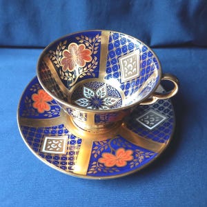 Puede incluir: Un juego de taza y platillo decorativo. La taza y el platillo son de color azul marino con detalles dorados y diseños florales en naranja y blanco. La taza tiene un asa dorada y un borde dorado. El platillo tiene un diseño a juego.