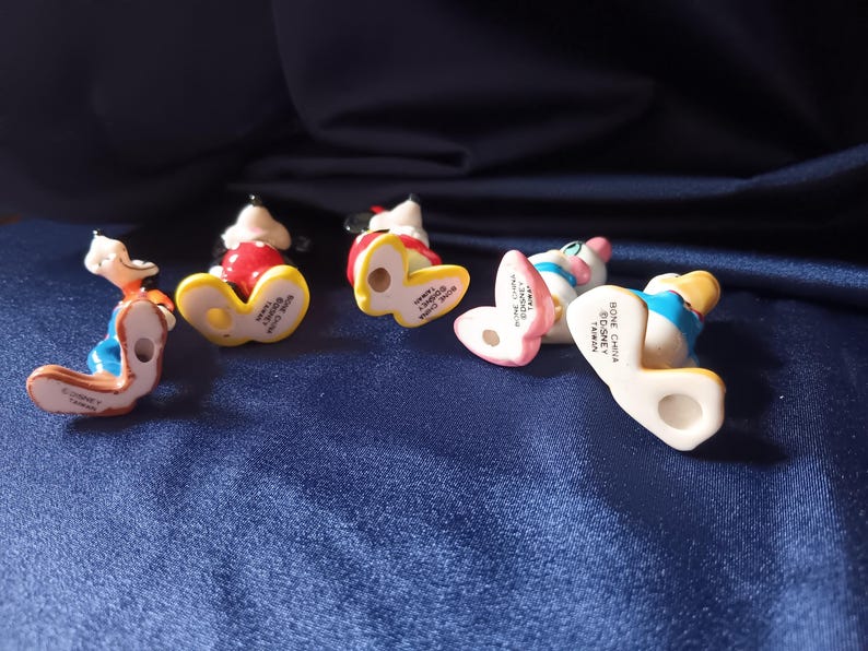 May include: Five small Disney figurines, including Mickey Mouse, Donald Duck, and Minnie Mouse, are arranged on a blue surface. The figurines are made of bone china and are in various poses, with their undersides visible.