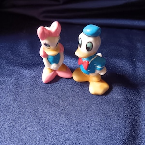 May include: Two ceramic figurines of Disney characters, Donald Duck and Daisy Duck. Donald Duck is wearing a blue sailor suit, a red bow tie, and a blue hat. Daisy Duck is wearing a pink bow, a blue top, and pink shoes. The figurines are on a blue surface.