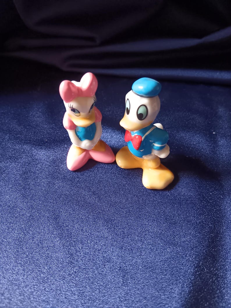 May include: Two ceramic figurines of Disney characters, Donald Duck and Daisy Duck. Donald Duck is wearing a blue sailor suit, a red bow tie, and a blue hat. Daisy Duck is wearing a pink bow, a blue top, and pink shoes. The figurines are on a blue surface.