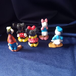 May include: A collection of small, colorful figurines featuring classic Disney characters. The figurines include Mickey Mouse, Minnie Mouse, Donald Duck, Daisy Duck, and Goofy. Each figure is painted with bright colors and stands on a small base.