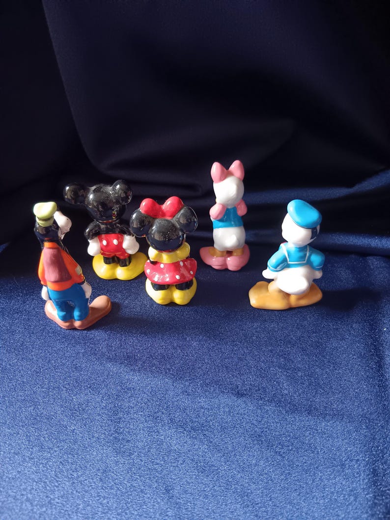 May include: A collection of small, colorful figurines featuring classic Disney characters. The figurines include Mickey Mouse, Minnie Mouse, Donald Duck, Daisy Duck, and Goofy. Each figure is painted with bright colors and stands on a small base.