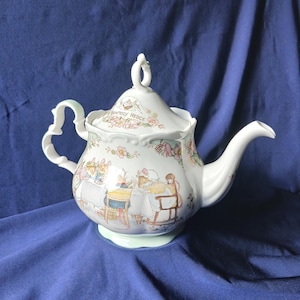 Puede incluir: Una tetera de cerámica blanca con tapa, asa y boquilla. La tetera presenta una base verde claro y está decorada con ilustraciones florales y de cuentos. La tapa tiene el texto "Brambly Hedge" y un asa de bucle.