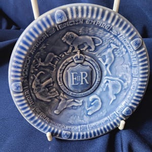 May include: A blue and white ceramic plate with a royal crest featuring the letters "ER" surrounded by animals representing different countries of the British Empire. The plate is inscribed with the words "British Empire" and the names of the countries represented.