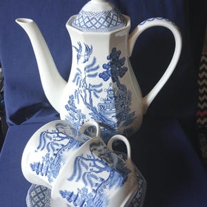 May include: A white ceramic teapot with a blue willow pattern featuring pagodas, willow trees, and birds. The set includes two matching cups and a saucer, all decorated with the same blue and white oriental-inspired landscape design.