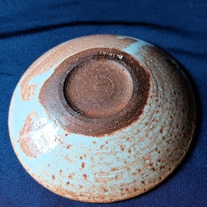 May include: A handmade ceramic bowl with a light blue and brown speckled glaze. The bowl's underside reveals a dark brown, textured surface with a central recessed area. The rim has a light blue glaze with a speckled pattern.