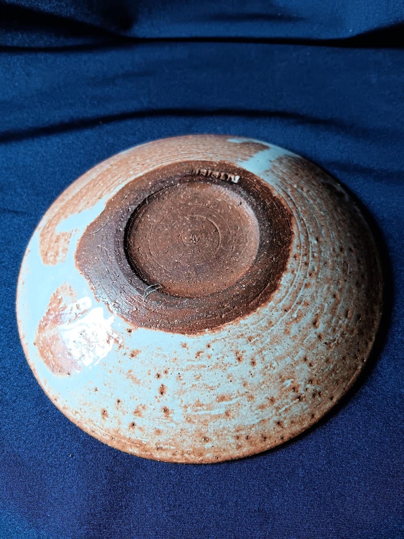 May include: A handmade ceramic bowl with a light blue and brown speckled glaze. The bowl's underside reveals a dark brown, textured surface with a central recessed area. The rim has a light blue glaze with a speckled pattern.