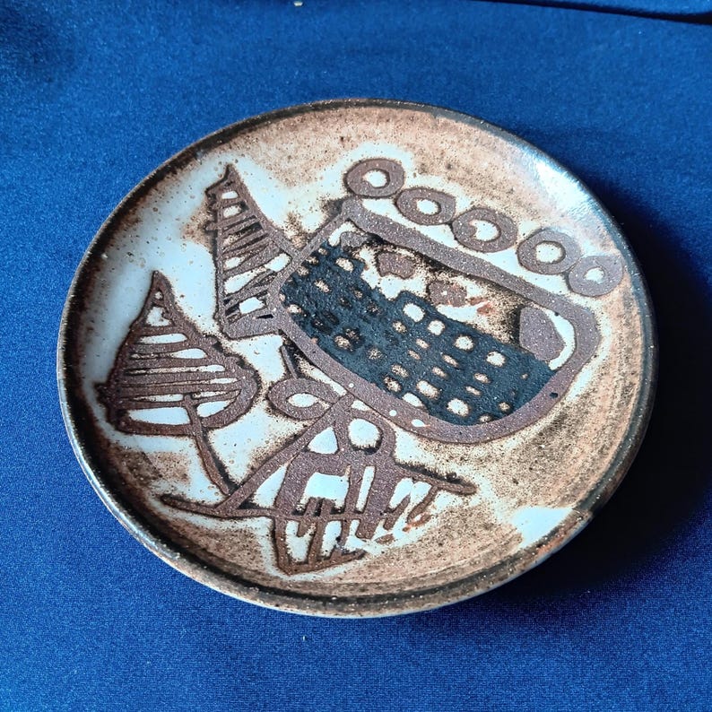 May include: A small, round ceramic plate with a brown and white abstract design. The plate features a central dark brown shape with a dotted texture, surrounded by other brown and white shapes. The rim is a lighter brown color.