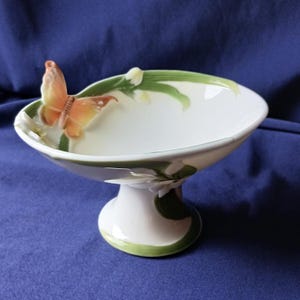 May include: A white ceramic pedestal bowl with a butterfly and floral design. The bowl has a rounded shape with a green and white rim, and a butterfly with orange and brown wings is perched on the side. The base is white with green accents.