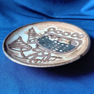 May include: A small, round ceramic plate with a brown and white abstract design. The plate features a central dark blue shape with a dotted pattern, surrounded by brown lines and shapes resembling trees and circles. The plate is on a blue surface.