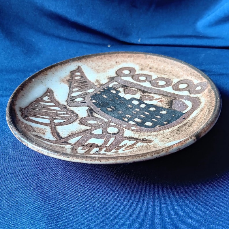 May include: A small, round ceramic plate with a brown and white abstract design. The plate features a central dark blue shape with a dotted pattern, surrounded by brown lines and shapes resembling trees and circles. The plate is on a blue surface.