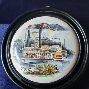 May include: A circular decorative plaque with a black frame featuring a colorful illustration of the Natchez steamboat. The ship has two smokestacks emitting dark smoke, with text reading Natchez, New Orleans, and U.S. Mail Packet visible.