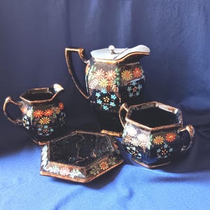 May include: A black ceramic tea set with floral designs in orange, yellow, and blue. The set includes a teapot, creamer, sugar bowl, and a small tray, all with gold trim.