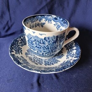 May include: A blue and white teacup and saucer set. The cup and saucer feature a detailed landscape design with floral accents. The set is made of ceramic and is placed on a blue surface. The teacup has a curved handle.