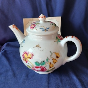 May include: A white ceramic teapot with a lid, spout, and handle. The teapot is decorated with colourful floral and insect designs in red, yellow, green, and blue. The teapot is sitting on a blue surface.