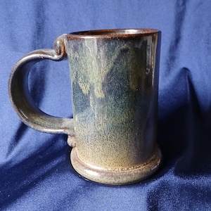 May include: A handcrafted ceramic mug with a dark brown rim and handle. The mug features a speckled, textured surface with shades of blue, green, and tan. The handle is a curved, sturdy design, and the base is a solid, rounded shape.
