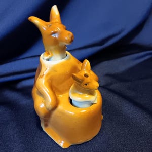 Puede incluir: Un juego de salero y pimentero de cerámica con una madre canguro y su cría. La madre canguro está sentada en una silla con su cría sentada en su regazo. Ambos canguros son marrones y tienen ojos negros.