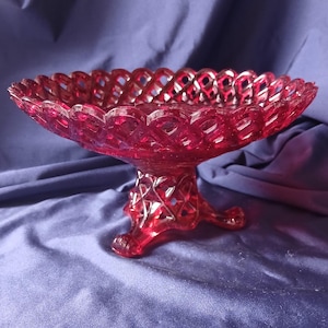 May include: A ruby red, vintage glass compote dish. The dish has a lattice design around the rim and on the pedestal base. The footed base has three small feet. The dish is displayed on a blue fabric surface.