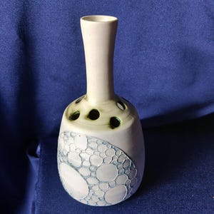 May include: A ceramic vase with a light beige neck and a bulbous body. The body features a textured, bubble-like pattern in shades of blue and white. The vase has several dark green holes near the neck.