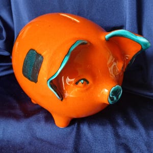 Puede incluir: Una hucha de cerámica naranja brillante con una ranura para monedas en la parte superior. El cerdo tiene detalles en color verde azulado en los bordes de las orejas, alrededor del hocico y una pequeña mancha rectangular en el costado.