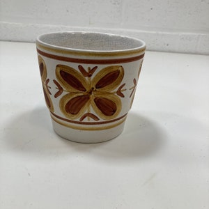Può includere: Un vaso da fiori in ceramica bianca con disegni floreali marroni. Il vaso ha un aspetto leggermente usurato e una forma arrotondata.