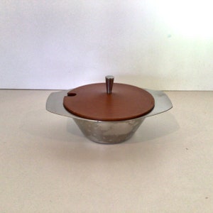 May include: A silver serving dish with a brown wooden lid and a silver knob. The dish has a rounded shape and a small spout on the side.