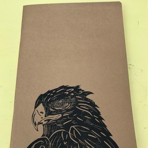 Peut inclure: Un carnet marron avec une estampe linogravure en noir et blanc d'une tête d'aigle sur la couverture.