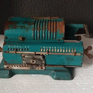 Puede incluir: Calculadora mecánica vintage de color turquesa, con signos visibles de óxido y desgaste. Cuenta con una serie de diales numéricos, una manivela y botones. La calculadora es probablemente de mediados del siglo XX.