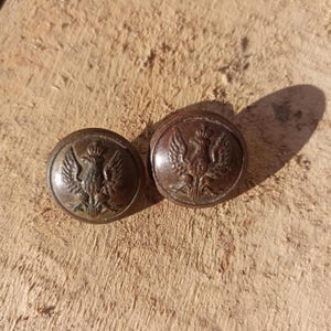 Puede incluir: Dos botones redondos de metal marrón, cada uno con un diseño en relieve de un águila con corona. El águila está representada con las alas extendidas y centrada en la cara de cada botón, que tiene una superficie lisa y ligeramente abombada.
