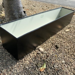 May include: A black metal planter box with a white interior. The planter is sitting on a bed of gravel.