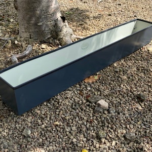 May include: A long, rectangular planter made of metal. The planter is dark blue with a white interior. It is sitting on a bed of gravel.