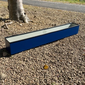 May include: A long, rectangular planter made of metal with a white interior and a dark blue exterior. The planter has a black rim and is sitting on a bed of small, grey stones.
