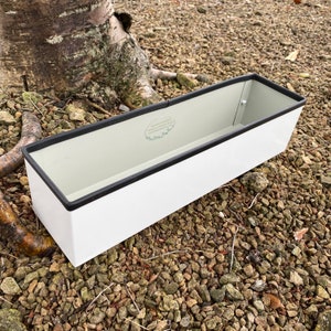 May include: White metal planter with a black rim. The planter has a small green logo on the side that reads "English Gardens Nursery & Greenhouse Supply".