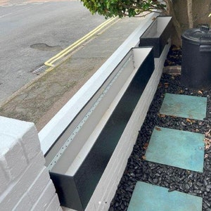 May include: Three rectangular black planters are mounted on a white brick wall. The planters have a black exterior and a white interior. The wall is next to a pavement and a bed of black pebbles with square stepping stones.