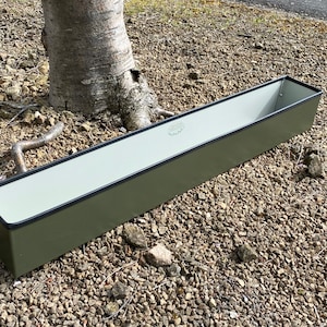 May include: A long, rectangular planter made of metal with a dark green exterior and a light grey interior. The planter has a black rim and a small white label with the text "The Planter Co."