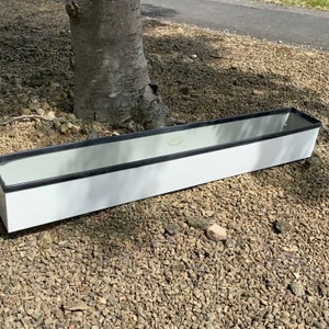 May include: A long, rectangular white planter box with a black trim. The planter is designed for plants and is set on a bed of gravel. The dimensions are suitable for a variety of plants. The planter is made of a durable material.