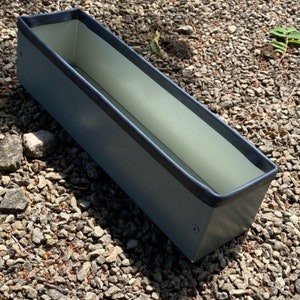 May include: A dark grey metal planter with a black rim. The planter is empty and sitting on a bed of gravel.