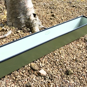 May include: A long, rectangular planter made of metal. The planter is painted a dark green colour with a light green interior. The planter is sitting on a bed of gravel.