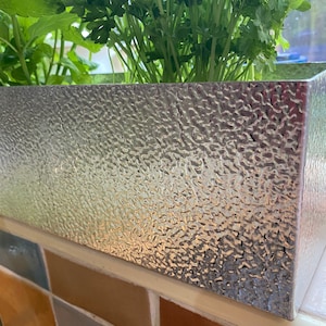 May include: A silver metal planter with a textured surface. The planter is filled with green leafy plants.