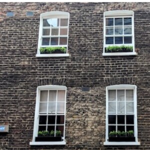 May include: A brick building with four white framed windows. Each window has a small planter box with green plants.