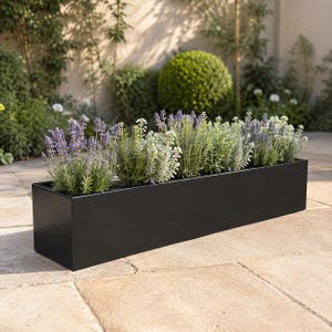 May include: A long, rectangular black planter box filled with a variety of green plants and purple lavender flowers. The container has a smooth, matte finish and a clean, linear design, holding multiple stems of foliage and blooming flowers.