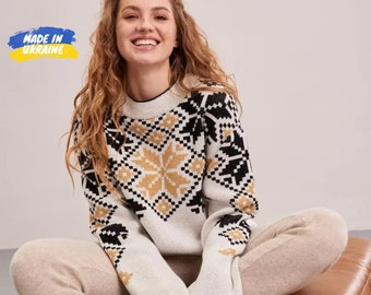 Pode incluir: Camisola branca com um padrão de floco de neve preto e dourado. A camisola tem mangas compridas e gola redonda. "Made in Ukraine" está escrito em azul e amarelo sobre um fundo branco.