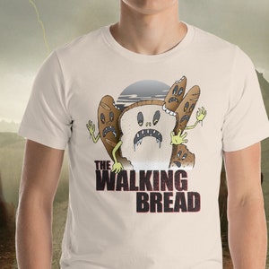 Puede incluir: Una camiseta blanca con un gráfico de una rebanada de pan con una cara y brazos, rodeada de otras rebanadas de pan con caras. El texto "The Walking Bread" está impreso debajo del gráfico.
