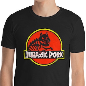 Peut inclure: T-shirt noir avec un logo Jurassic Park rouge et jaune représentant un squelette de cochon de dessin animé avec le texte "Jurassic Pork".