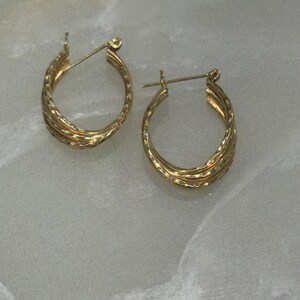 May include: A pair of gold hoop earrings featuring a triple-layered design with a textured, twisted finish. Each earring is constructed with a hinged post closure, showcasing a polished metallic surface that reflects light across the curved bands.