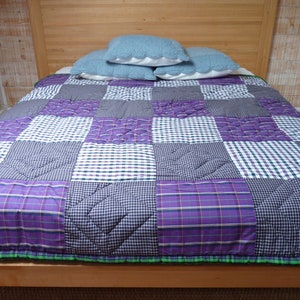 Op de afbeelding: Een patchworkdeken met een paars, wit en blauw geruit patroon. De deken is gemaakt van verschillende stoffen en heeft een gestikt ontwerp.