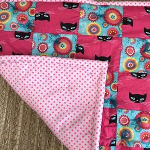 May include: A pink and blue patchwork quilt with a cat and flower design. The quilt is made of squares of different fabrics, each with a different pattern. The quilt is folded in half and the back side is pink with white polka dots.